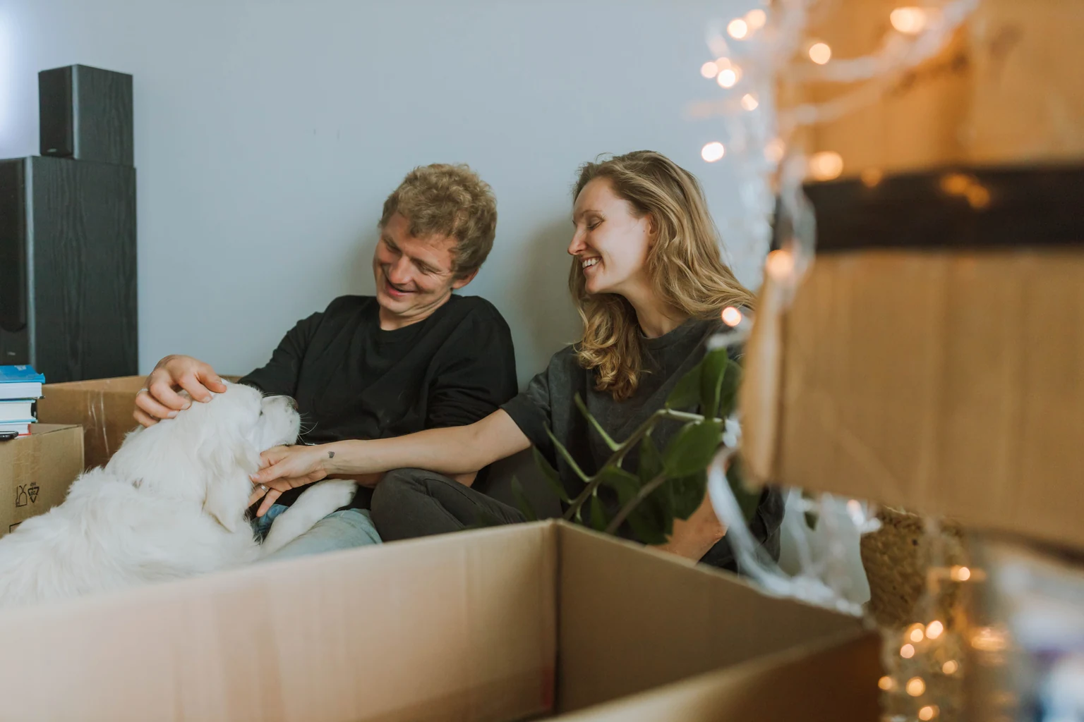 A couple petting their white dog while sitting among moving boxes in their new home